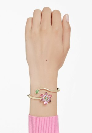Gold bracelet featuring a pink flower with green accents and a curved design. Worn on a hand, with a ribbed pink sweater visible below.