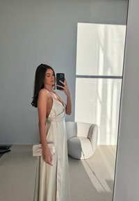 Crèmezijde spaghetti strap jurk met een diepe V-hals en een gedrapeerde voorkant. Houdt een gestructureerde clutch vast, staande naast een gebogen stoel.