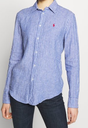 Femme portant une chemise rayée verticale bleu et blanc avec un petit logo rouge brodé sur le côté gauche de la poitrine.