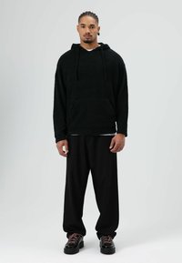 Schwarzer Fleece-Hoodie mit Fronttasche, kombiniert mit lockeren schwarzen Hosen und klobigen schwarzen Schuhen mit pinken Schnürsenkeln. Schlichter Hintergrund.