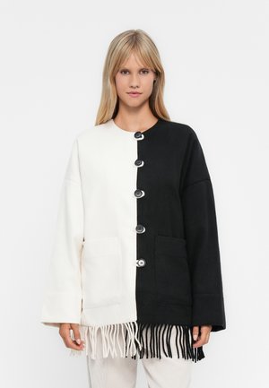 Woman wearing a black and white two-tone buttoned jacket with front pockets and fringe at the hem, standing against a plain background.