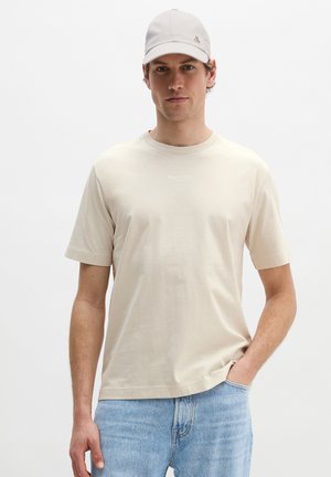 Man met beige pet en beige T-shirt met korte mouwen en subtiele tekst, lichtblauwe spijkerbroek, staand tegen een effen witte achtergrond.