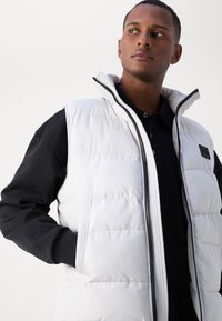 Gilet en duvet blanc avec un col haut, présentant des poches latérales et des accents noirs sur les épaules, sur un polo noir. Texture lisse.