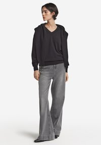 Czarna, oversize bluza z kapturem, z dekoltem w kształcie litery V i sznurkiem do regulacji, zestawiona z szarymi spodniami dzwonami o wysokim stanie. Gładka tekstura, nowoczesny krój i bogaty kontrast kolorów.