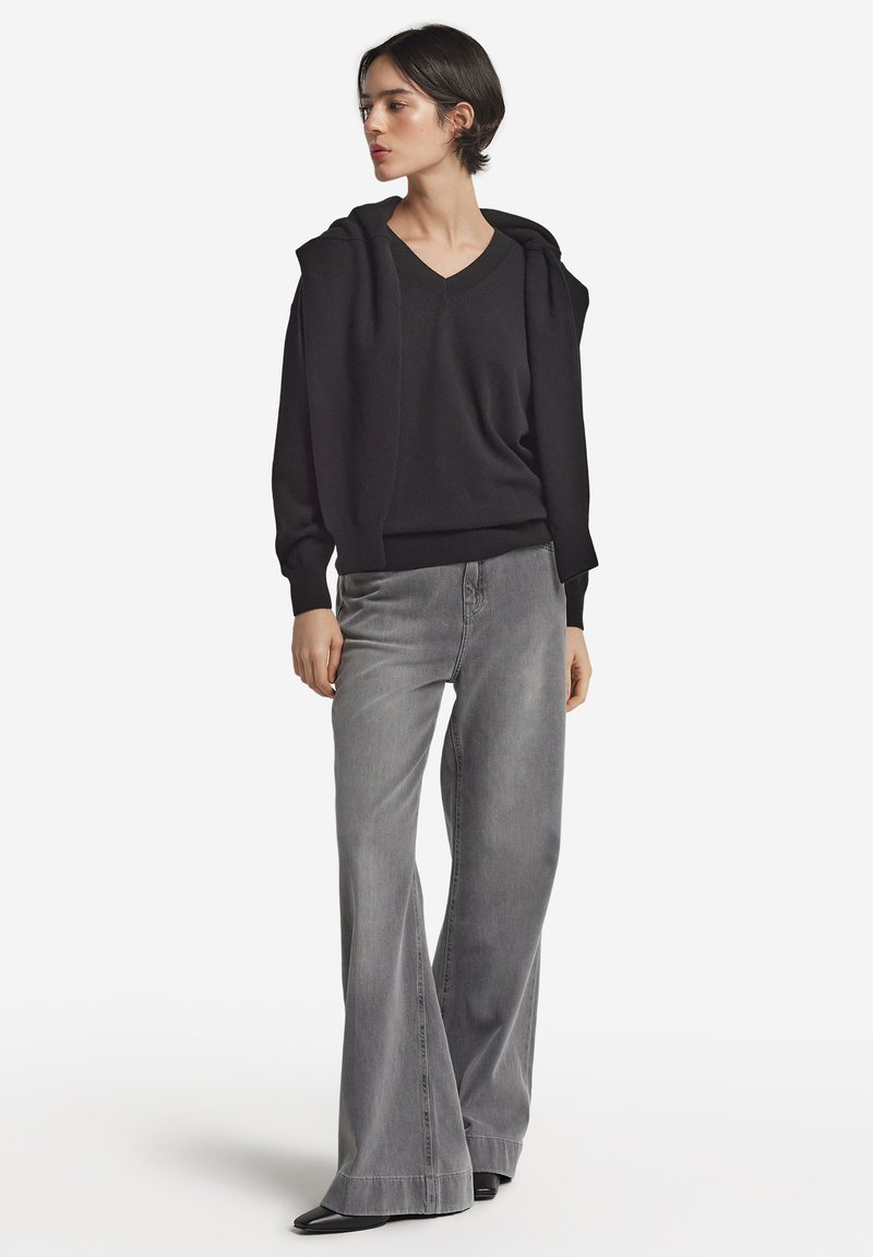Czarna, oversize bluza z kapturem, z dekoltem w kształcie litery V i sznurkiem do regulacji, zestawiona z szarymi spodniami dzwonami o wysokim stanie. Gładka tekstura, nowoczesny krój i bogaty kontrast kolorów.
