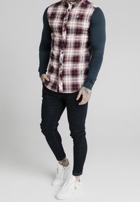 Camicia a maniche button-up con motivo a quadri rosso, nero e bianco, abbinata a jeans scuri aderenti e sneaker bianche. Sotto, una camicia blu a maniche lunghe.