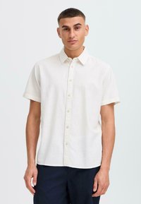 Camisa de botón de manga corta de color crema claro, hecha de tejido texturizado, con cuello, botones en la parte frontal y mangas enrolladas.