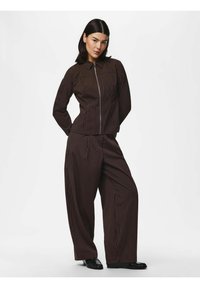 Femme aux cheveux foncés portant une veste marron foncé à fines rayures avec fermeture éclair et un pantalon large assorti avec des chaussures noires sur fond blanc.