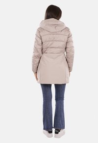 Cappotto puffo beige chiaro con cappuccio, caratterizzato da un design texturizzato e trapuntato e vita cinchata. Abbinato a jeans svasati e scarpe da ginnastica.