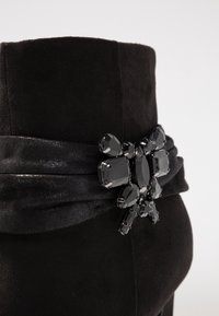 Botín de ante negro que presenta un detalle de tela envolvente y un adorno de joya con piedras negras de diversas formas en la parte trasera.