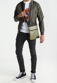 Mężczyzna w ciemnozielonej kurtce bomber, graficznej białej koszulce, czarnych dopasowanych dżinsach, czarnych sneakersach za kostkę oraz oliwkowej torbie na ramię.
