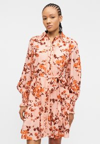 Γυναίκα με πλεξούδες, ντυμένη με ροζ φόρεμα με λουλουδάτο μοτίβο, μακριά φουσκωτά μανίκια και ζώνη στη μέση, στέκεται μπροστά σε απλό φόντο.