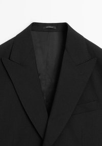 Zwarte op maat gemaakte blazer met een geknipte revers, enkele borstzak en een gladde stoftextuur. Verzorgingslabel zichtbaar aan de binnenkant van de kraag.