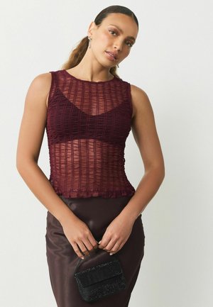 Burgunderfarbene Bralette-Oberteil aus strukturiertem Stoff, mit durchsichtigen Einsätzen und gerafftem Saum, kombiniert mit einem braunen Satinrock und einer kleinen schwarzen Handtasche.