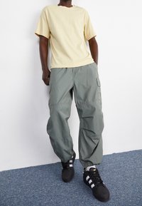 Světlě žluté tričko s krátkým rukávem, volné zelené cargo kalhoty s bočními kapsami a černé tenisky s bílými pruhy. Hladká textura látky.