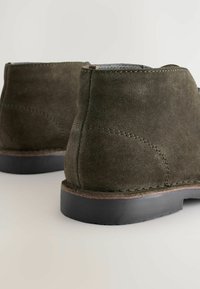 Botines de ante en verde olivo oscuro con una textura suave, con costuras en contraste y una suela de goma negra para mayor tracción.