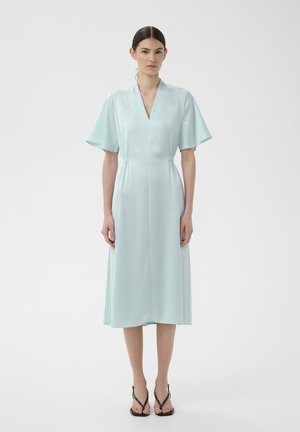 Naine seisab seljas kahvatu sinise V-kaelusega midi-pikkuses kleit lühikeste lahtiste varrukatega ja mustade kontsadega sandalites tavalise heleda tausta ees.