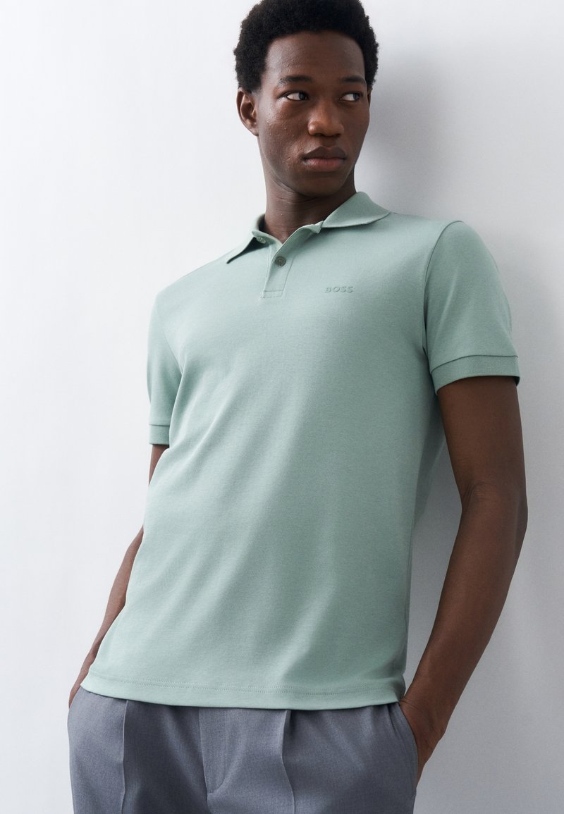 Νέος άνδρας με ανοιχτό πράσινο πόλο μπλούζα και γκρι παντελόνι, στέκεται με τα χέρια στις τσέπες, κοιτώντας προς το πλάι μπροστά σε λευκό τοίχο.