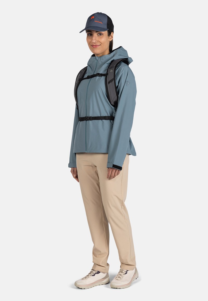 Chaqueta impermeable azul claro con capucha, herrajes negros y cinturón a la cintura, combinada con pantalones beige y zapatos de senderismo de color claro.