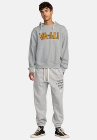 Szary sweatshirt z napisem "VAALI" w pomarańczowym kolorze, zestawiony z szarymi spodniami dresowymi z tekstem, elastycznymi mankietami i białymi sznurkami. Czarne trampki.