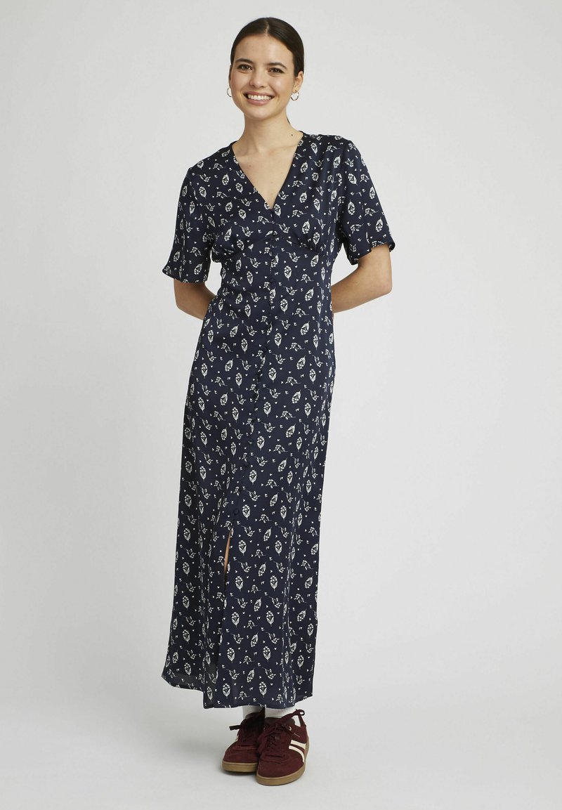 Robe maxi florale bleu marine avec un décolleté en V, des manches courtes et une fente sur le côté, associée à des baskets bordeaux et des chaussettes blanches.