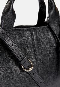 Černá texturovaná kožená kabelka s nastavitelným popruhem a zlatou přezkou, zobrazena zblízka na bílém pozadí.