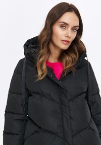 Czarna kurtka puchowa z kapturem, z pikowanym wzorem, z zapięciem na zamek z przodu i regulowanymi sznurkami, noszona na różowej bluzce.