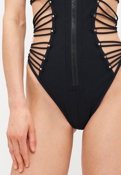 Black swimsuit with a zipper closure and cut-out sides, featuring thin black straps and circular gold accents along the openings.