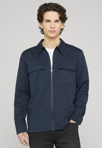 Veste zippée bleu marine avec col, deux poches poitrine et manches longues. Confectionnée dans un tissu lisse de poids moyen.