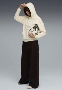 Donna in felpa crema e pantaloni neri larghi con borsa bianca Puma, che sistema il cappuccio su uno sfondo grigio.