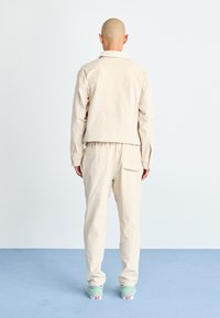 Beige, lätt jacka med krage och avslappnad passform, tillsammans med matchande byxor. Outfiten har en ren design och neutrala färger.