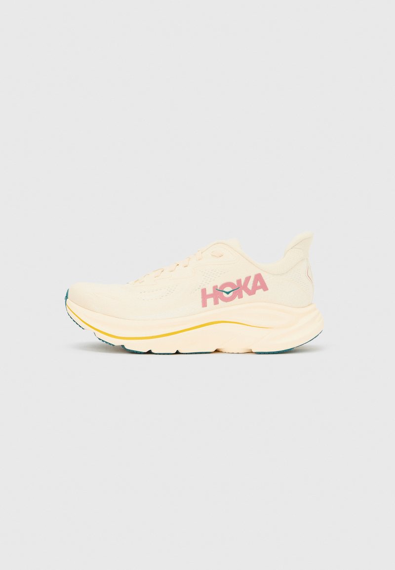 Μπεζ αθλητικό παπούτσι με υφασμάτινο επάνω μέρος, που φέρει ροζ λογότυπο "HOKA", στρογγυλή μύτη, ενδιάμεση σόλα με κίτρινες λεπτομέρειες και λαστιχένια εξωτερική σόλα.