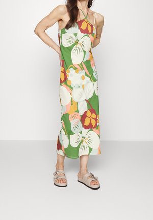 Mujer con un vestido verde sin mangas hasta el tobillo con grandes estampados florales de colores y sandalias planas beige con correas de hebilla.