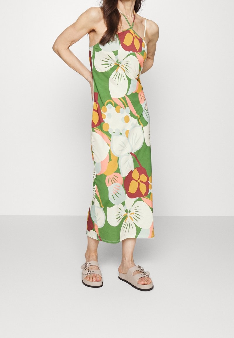 Femme portant une robe verte sans manches, longueur cheville, avec de grands motifs floraux colorés et des sandales plates beiges à brides bouclées.