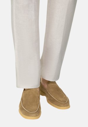 Pantalon en lin beige clair porté avec des mocassins à enfiler en daim marron clair, avec des coutures apparentes et des semelles en caoutchouc.