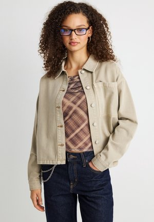 Νέα γυναίκα με σγουρά μαλλιά που φορά γυαλιά μπλε φωτός, μπεζ κοντό μπουφάν, καρό τοπ, σκούρα τζιν με αλυσίδα, ποζάρει μπροστά σε λευκό φόντο.