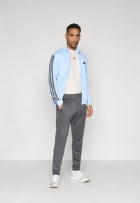 Casaco azul claro com fecho éclair e riscas cinzentas, t-shirt branca com detalhes vermelhos, calças de fato de treino cinzentas e sapatilhas desportivas brancas. Design simples.