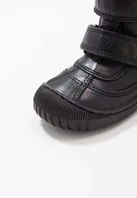 Scarpa nera in pelle con due cinturini in Velcro, caviglia imbottita, punta arrotondata e suola in gomma testurizzata con scanalature per una migliore trazione.