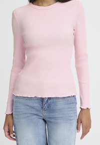 Sieviete, tērpusies pielāgotā gaiši rozā ribotu garām piedurknēm blūzē ar viļņotām apakšmalām un gaiši zilās augsto jostasvietu džinsos, stāv pretī baltajai fona.