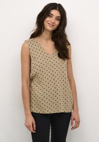 Top sans manches beige avec décolleté en V, orné de pois noirs, avec une coupe ample et une texture de tissu lisse. Porté avec un pantalon en denim foncé.