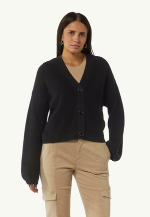 Frau in einem schwarzen, geknöpften Cardigan über einem beigen Oberteil und beigen Cargo-Hosen, die mit einer Hand in der Tasche vor einem schlichten Hintergrund steht.