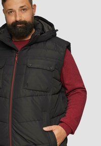 Schwarzige gepolsterte Weste mit Kapuze, zwei Brusttaschen und roten Reißverschlussdetails. Getragen über einem roten strukturierten Langarmshirt.