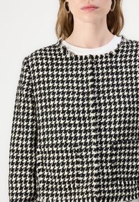 Jachetă în motive houndstooth alb-negru, țesătură texturată, marginile desfăcute, guler rotund, două buzunare frontale și croi ajustat.