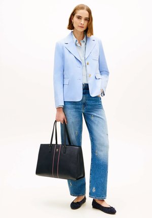 Woman wearing light blue blazer and jeans with embroidered logos, holding a large black handbag, standing against a white background.