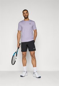 T-shirt lilás de manga curta com o logo EA7, combinado com calções pretos com o logo. Segura uma raquete de ténis, vestindo sapatilhas e meias brancas desportivas.