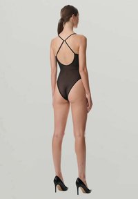 Kvinna som står i en svart genomskinlig bodysuit med korslagda ryggband och svarta högklackade skor mot en grå bakgrund.