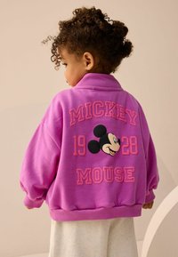 Růžová oversized mikina s nápadnými rukávy, textem "MICKEY MOUSE", detailem "1928" a grafikou Mickey Mouse na zadní straně.
