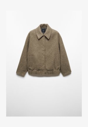 Brun uldtekstureret jakke med trykknapper over en hvid sweater, kombineret med mørkegrå bukser, en hånd i lommen.