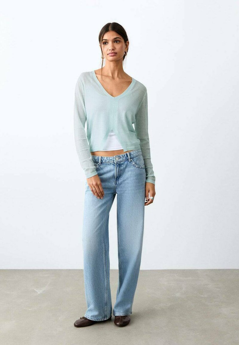 Donna che indossa un cardigan corto azzurro chiaro sopra una maglietta bianca, jeans larghi azzurri chiari e scarpe marrone scuro, in piedi contro una parete bianca liscia.