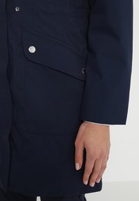 En marinblå vattentät jacka har en sidoficka med en metallknäppning och en mjuk tygkänsla, tillsammans med en rak ärm.